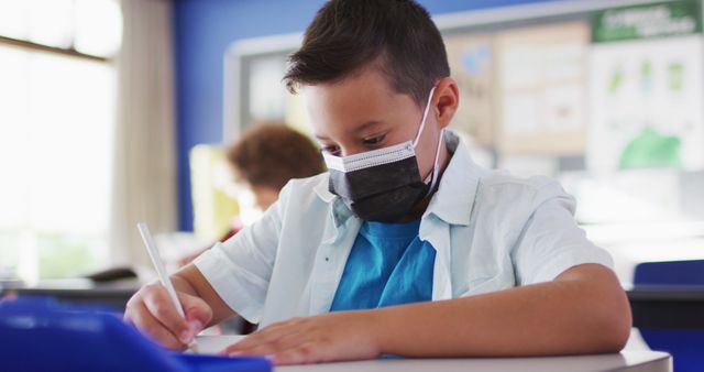 Portrait of biracial schoolboy wearing face mask in classroom looking at camera. children in primary school during coronavirus covid 19 pandemic.