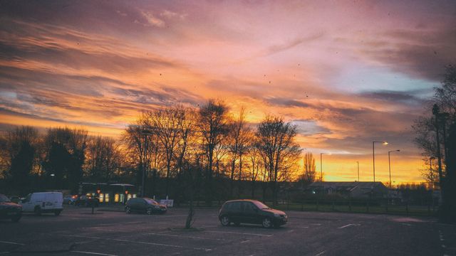 Vibrant, dramatic sunset over an urban parking lot. Trees are silhouetted against an orange and pink sky. A few cars are scattered around. Ideal for themes of tranquility in urban settings, end-of-day photography, city life, and nature in the city.