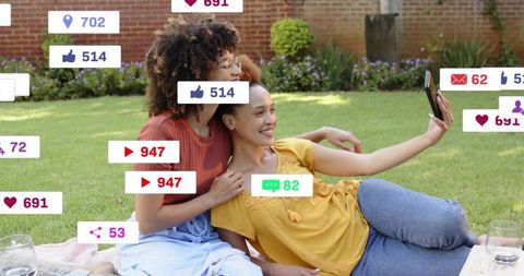 Two women taking selfie on picnic blanket in sunlit backyard with social media icons