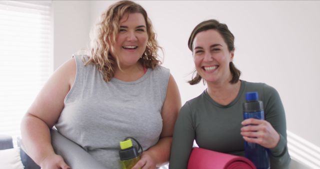 Two women smiling after completing yoga class, one holding yellow water bottle and gray mat, the other holding blue water bottle and pink mat. Indoors with natural light from the window. Perfect for promoting fitness and wellness programs, healthy lifestyle, yoga classes, and women's health initiatives.