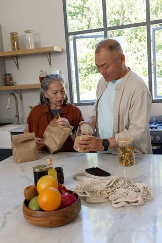 Senior asian couple packing chickpeas and pasta on sunlit marble kitchen island