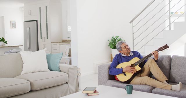 Elderly man is playing an acoustic guitar while sitting on a sofa in a modern and bright living room. There is a cup and books on the coffee table in front of him. This image can be used to illustrate concepts of leisure, music, relaxation, and enjoying retirement. Ideal for articles, blogs, or advertisements related to senior lifestyle, home living, and hobbies.