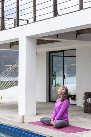Mature woman meditating at poolside terrace wearing headphones on purple yoga mat