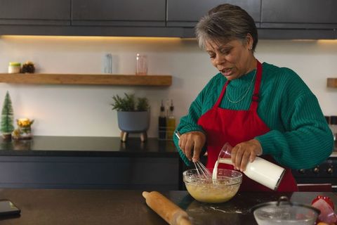 Mature woman pouring milk and whisking batter in cozy kitchen wearing red apron