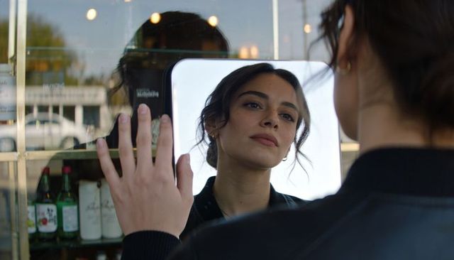 Young woman gazing at reflection in handheld mirror against shop window with bottles