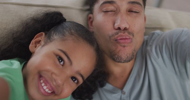Father and daughter sitting on couch taking a selfie and making funny faces creates a warm family moment. Ideal for use in family-focused marketing, parenting blogs, social media posts about father-daughter relationships, and advertisements promoting family togetherness.