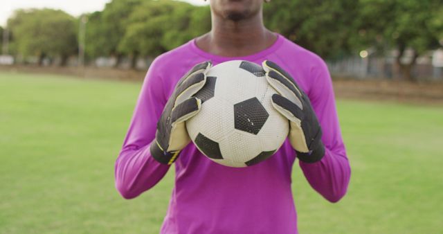 Depicting a young soccer player in action holding a ball with both hands while wearing a pinkish-purple jersey and gloves on a lush green field. Use this for content related to soccer training, youth sports, football clubs, athleticism, and fitness programs.