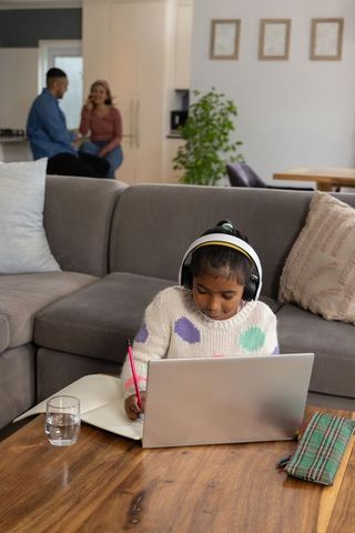 Asian girl studying on laptop with headphones in cozy living room while parents talking nearby