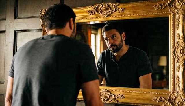 Contemplative man leaning and examining reflection in ornate gilded mirror in paneled study