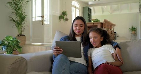 Mother and daughter sharing tablet on sofa, enjoying screen time in bright open-plan home