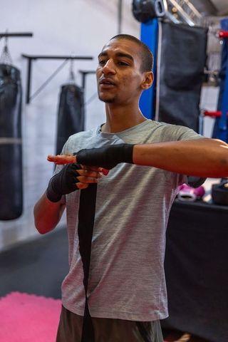 Focused young boxer wrapping hands before training in gym with heavy bags and ring