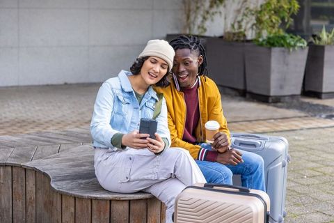 Multicultural young travelers sharing smartphone and coffee with suitcases on city bench
