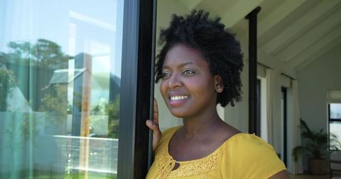 African american woman standing at sliding glass door smiling in modern sunlit home