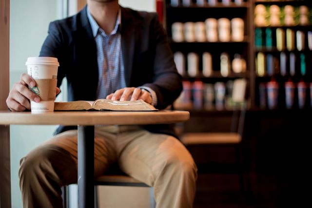 A man in a casual blazer and khakis enjoys a coffee while reading a book in a modern cafe with a background of assorted merchandise on shelves. Perfect for concepts related to relaxation, leisure time, studying in a coffee shop, and modern lifestyle scenes.