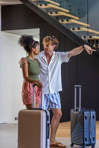 Diverse couple arriving with suitcases in modern rental foyer under floating glass staircase