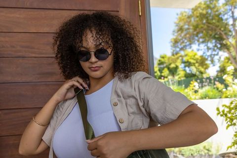 Curly-haired woman adjusting olive bag strap wearing sunglasses and beige overshirt on sunlit porch