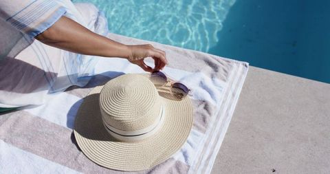 African american woman reaching for sunglasses beside straw hat on sunlit pool deck towel