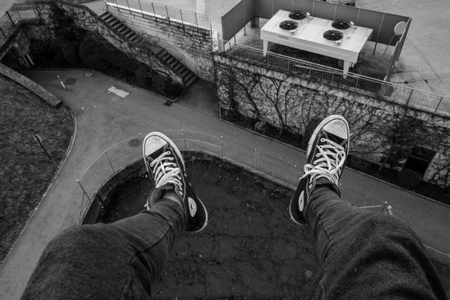 This image showing feet dangling over a height in black and white is ideal for themes of risk-taking, adventure, urban exploration, and perspective. Suitable for use in articles, blogs, and websites discussing thrill-seeking activities or urban photography.
