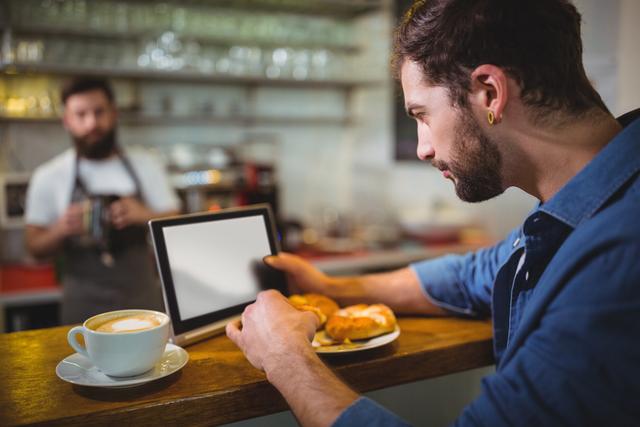 Man sitting at counter and using digital tablet while having croissant in cafÃ©