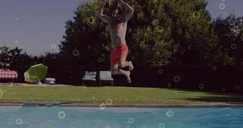 Man jumping into backyard pool in red swim trunks, picnic umbrella and lounge chairs