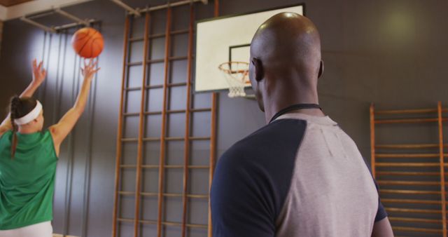 Coach watching basketball player taking shot in indoor gym. Useful for content related to sports training, coaching techniques, physical fitness, skill development, teamwork, athleticism, and educational settings.