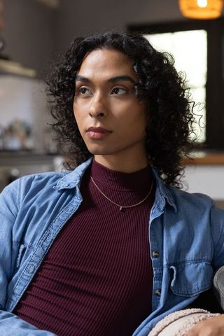 Non-binary young adult sitting on couch wearing denim shirt and layered gold necklaces