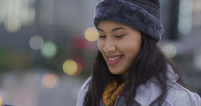 Young woman smiling while wearing warm winter clothes, including a hat and coat, in an urban setting. This image can be used to represent happiness during cold weather, winter fashion, or outdoor winter activities in a city environment.