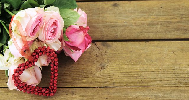 A bouquet of pink and white roses lies next to a red heart-shaped bead arrangement on a wooden surface, with copy space. Perfect for themes of love, Valentine's Day, or romantic celebrations.
