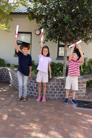 Three children holding diplomas and wearing caps in sunlit school courtyard celebration