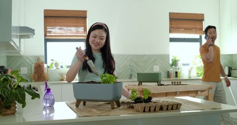 Woman planting indoor herb garden on sunlit kitchen island while partner talking on phone