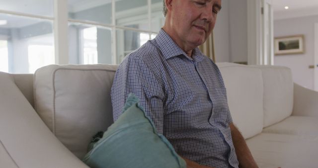 This image shows a senior man sitting comfortably on a couch, relaxing in a modern and well-lit living room. Ideal for use in advertisements, articles, or social media posts related to senior lifestyle, home comfort, retirement, and relaxation.