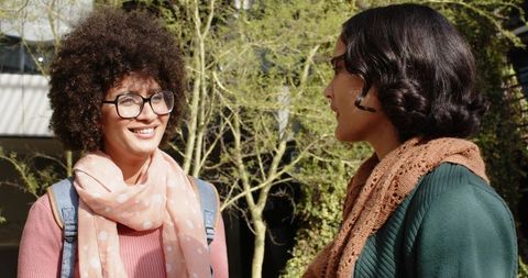 Smiling Female Friends Talking on Sunlit Campus Wearing Scarves, Sweaters and Backpacks