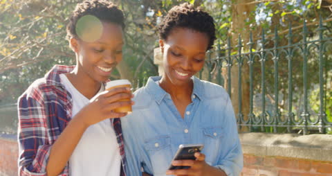Twin Sisters Smiling and Using Mobile Phone While Walking with Coffee