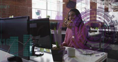 Woman in pink blouse eating while analyzing data on monitors in open-plan office