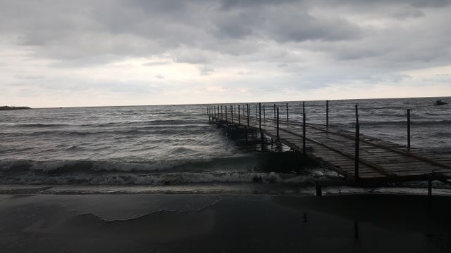 Wooden pier stretching over rough sea waves on an overcast day with a cloudy sky. Useful for illustrating seaside solitude, nature's power, melancholic coastal scenes, or themes of peace and tranquility.
