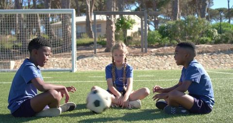 Diverse youth soccer teammates sitting on turf passing ball by goal during sunny practice