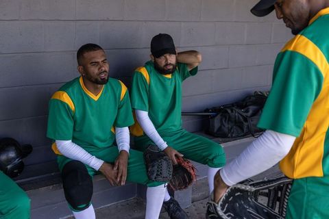 Baseball players resting in dugout between innings holding gloves and bats