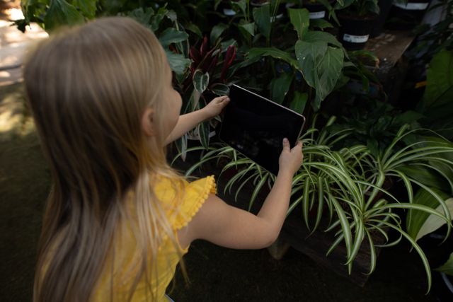 Caucasian girl with long blonde hair enjoying time in a sunny garden, exploring, holding digital tablet taking photos of plants. Gardening botany hobby.