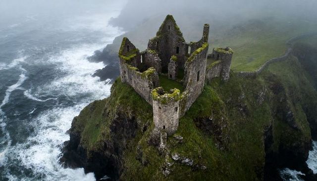 Misty cliffside castle ruins overlooking stormy sea, windswept promontory fortress