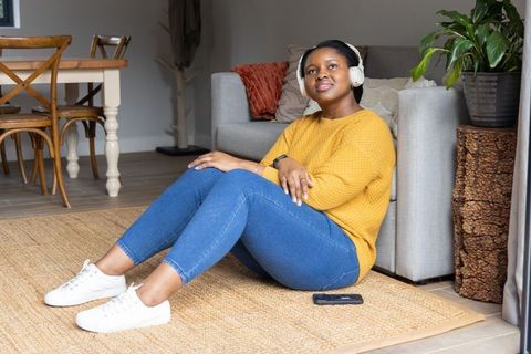 Relaxed african american woman listening on headphones at home sitting on rug beside sofa