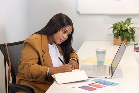 African american woman working at desk writing notes viewing laptop in minimalist office
