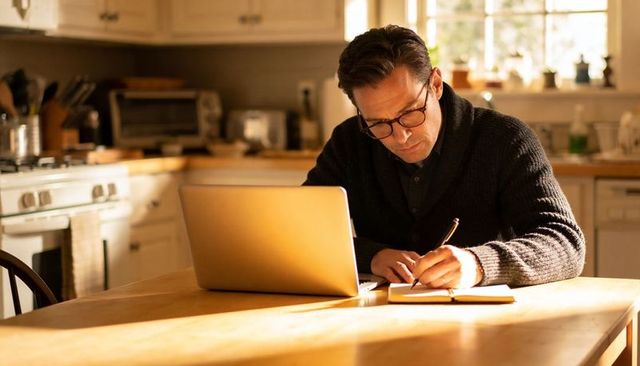 Sunlit kitchen workspace: middle-aged man writing notebook and using laptop on wooden table