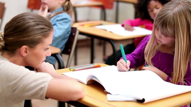 A teacher kneeling next to a young student, offering assistance with a workbook. The video showcases direct teacher-student engagement, emphasizing personalized education and instructional support in an elementary school setting. Ideal for educational content, blogs on teaching strategies, or promotional material for academic support programs.