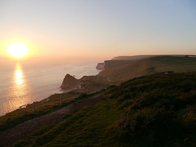 This image shows a stunning sunset over coastal cliffs at Durdle Door, with the sun sinking into the horizon and casting a warm glow over the sea. Ideal for use in travel brochures, landscape photography collections, and websites promoting natural beauty or outdoor activities.