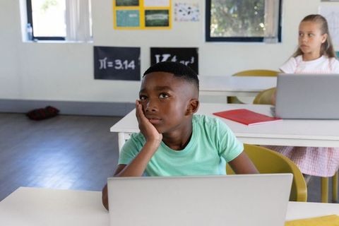African american boy daydreaming at desk with laptop in modern classroom learning