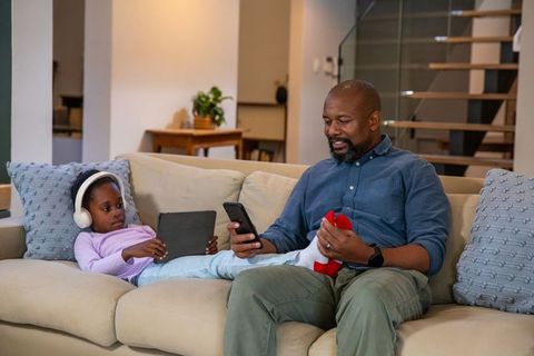 African american father bonding with daughter on couch sharing devices during cozy night
