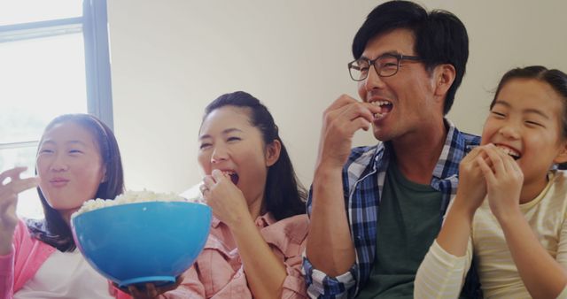 Family sitting on couch, sharing bowl of popcorn, smiling and enjoying time together. Perfect for themes of family bonding, leisure time, or joyful moments. Suitable for use in family-oriented advertisements, articles on family activities, or websites promoting togetherness.