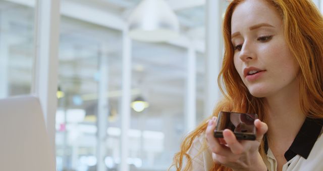 Young professional woman interacting with her smartphone’s voice assistant in an office environment. Ideal for applications of modern workspaces, technology in business, professional communication, and workflow enhancements.