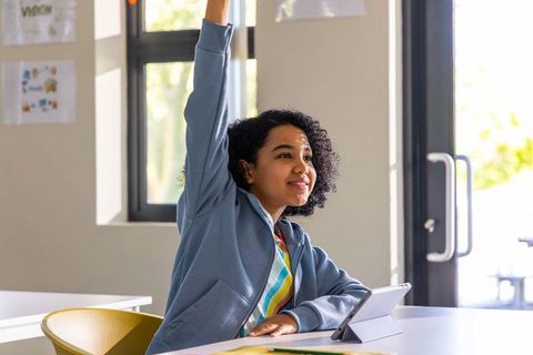 Student raising hand confidently in bright classroom using tablet for learning