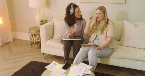 Two women collaborating on home budgeting with tablet and laptop on sofa with papers
